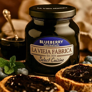 Jar of La Vieja Fabrica blueberry marmelade with slices of bread spread with it, surrounded by blueberries and mint leaves.